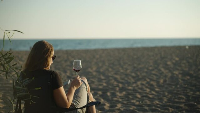 A girl on the beach by the sea photographs a glass of wine in her hand against the sunset. Picnic on the beach.