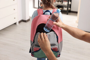 Father putting bottle of water into daughter`s backpack at home, closeup. Preparing to school