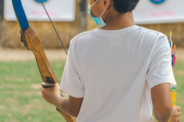 Back view of Asian boy wear face mask aims archery bow and arrow to colorful target in shooting...
