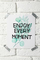 Card with phrase Enjoy Every Moment attached on white brick wall