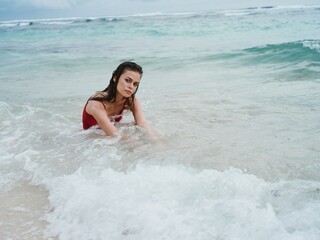 A woman in a red swimsuit sits on the sand by the ocean in the waves on the beach of Bali Island...