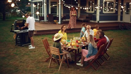 Group of Multiethnic Diverse People Having Fun, Communicating with Each Other and Eating Barbecue at Outdoors Dinner. Family and Friends Gathered Outside Their Home on Warm Summer Afternoon. - Powered by Adobe
