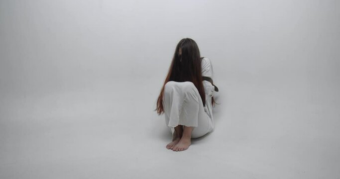 Cinematic footage of girl with face covered with  long red hair, dressed in straitjacket, barefoot,  tied with straps, sits in the corner of white room. Ward in mental hospital. Horror psycho concept