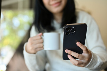 Pretty young Asian woman using her smartphone while sipping coffee in the morning at home.