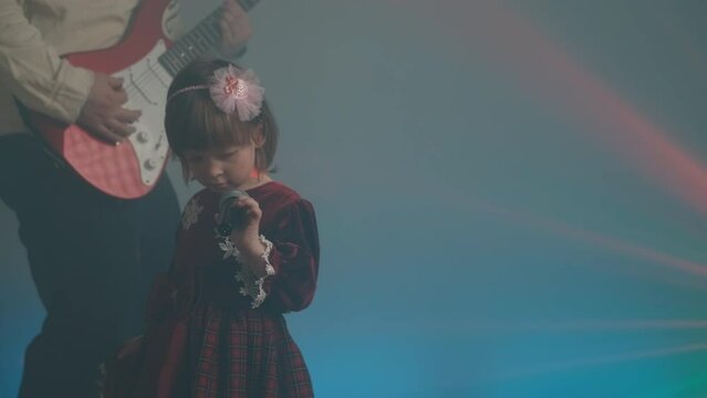 Little girl on stage in vintage dress, she sings into microphone and dances, her father plays an electric guitar. Color music is shining and smoke is billowing. Family time, creativity and hobbies