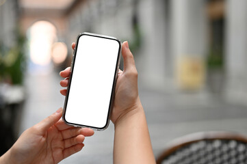 A smartphone white screen mockup is in a woman's hands over blurred background of city square.