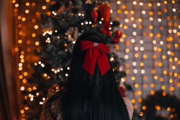 A brunette woman with a red bow in her hair sitting near the Christmas tree with festive decorations. New Year and Merry Christmas winter holiday celebration.