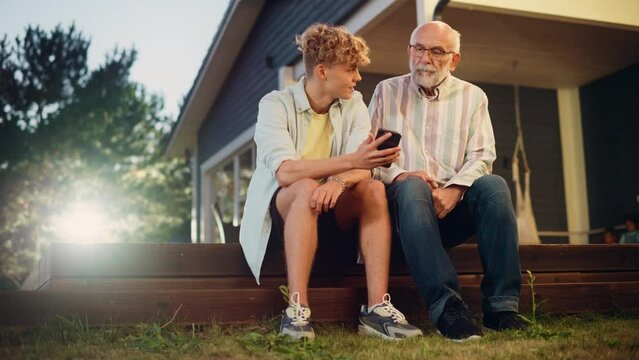 Handsome Teenage Grandson Teaching His Grandfather to Use a Smartphone. Young Man Showing Family Photos and Videos to His Grandparent. Relatives Sitting Outside on a Porch on a Nice Summer Day.