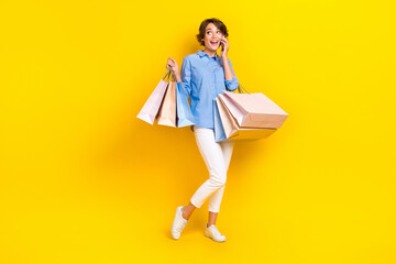 Full length photo of excited cute lady wear blue shirt holding shoppers talking device looking empty space isolated yellow color background