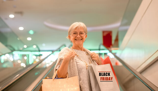 Attractive Smiling Older Woman Carries Shopping Bags As She Climbs The Escalator In A Shopping Mall. Black Friday And Consumerism Concept