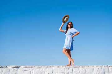 Lifestyle portrait of a happy young relaxed woman in a blue dress sitting on stone fence on the background of the blue summer sky. Summer vacation concept.