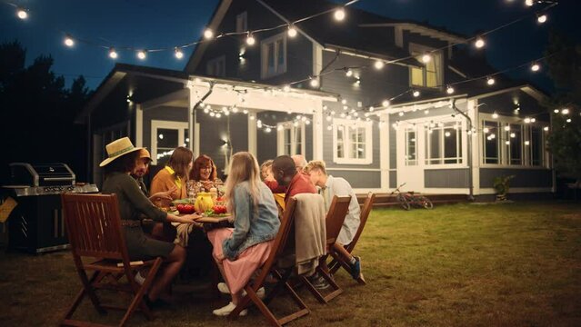 Parents, Children and Friends Gathered at a Barbecue Dinner Table Outside a Beautiful Home with Lights Decorations. Old and Young People Have Fun and Eat Food. Garden Party Celebration in a Backyard.