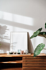 Modern minimalist Scandinavian style interior with white poster mockup, candles and tropical green home plants on a wooden console under sunlight and shadows on a gray wall. Selective focus
