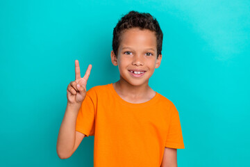 Photo of excited sweet little guy dressed orange t-shirt showing two v-signs isolated teal color...
