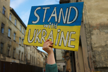 Young woman holding poster in colors of national flag and words Stand with Ukraine outdoors, closeup