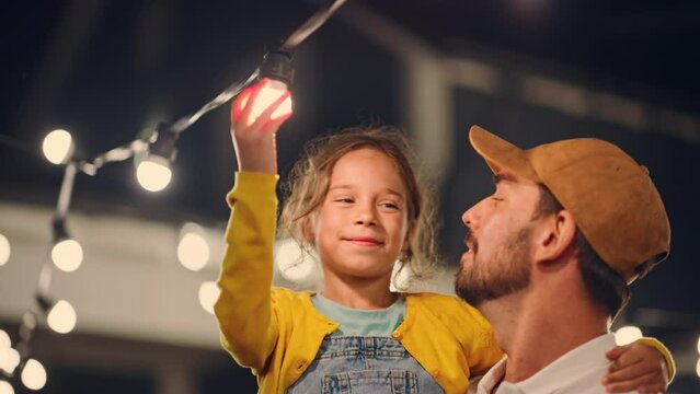 Cinematic Shot of a Father Holding His Small Daughter in His Arms, Helping Her to Fix a Lightbulb in a Backyard Lights Installation. Father and Daughter High Five and Celebrate Successful Repair.
