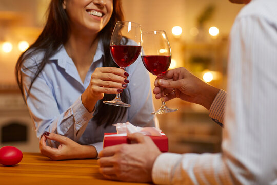 Happy couple enjoying a spontaneous romantic dinner at home. Husband and wife drinking red wine on Valentine's Day. Man in love gives a present to his beloved woman and raises a toast. Crop, close up