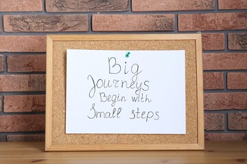 Corkboard and phrase Big Journeys Begin With Small Steps on table against brick wall. Motivational...