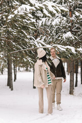 A couple in love spend time together walking in a snowy forest among trees having fun and laughing in nature on vacation. A young man and a woman in warm clothes travel outdoors. Selective focus