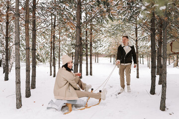 Fototapeta na wymiar A couple in love spend time together walking in a snowy forest among trees having fun and riding a sleigh in nature. A young man and a woman in warm clothes are traveling. Selective focus