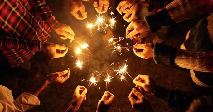 Handheld top view shot, Group of young friends stand in circle holding fire burning sparklers, They are raising and waving playing with sparkly in hands on outdoor new year's party night
