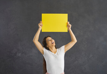 Happy young Latino woman on black studio background show yellow mockup paper sheet. Smiling millennial Hispanic female demonstrated mockup banner paperwork recommend sale deal or promotion.