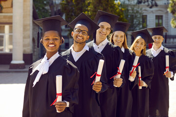 Group of happy diverse university graduates with diplomas. Six multiethnic students in graduation caps and gowns standing outside university building in campus yard, looking at camera and smiling