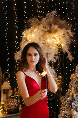 a girl in a red silk tight-fitting dress stands in a dark room against the background of New Year's decorations and holds a sparkler in her hands