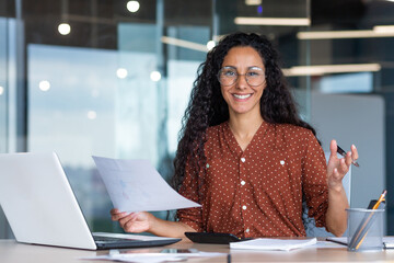 Fototapeta na wymiar Portrait of happy and successful hispanic woman, businesswoman smiling and looking at camera holding contracts and invoices, working inside office with laptop on paper work.
