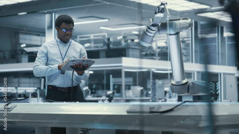Aufkleber Industrial Robotics Engineer Working on Robotic Arm in a High Tech Factory. Black Male Scientist Uses Tablet Computer to Manipulate and Program the Robot to Pick Up and Move a Metal Component.