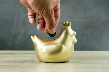 A man with a Thai coin carrying to chicken bank on wooden table and dark background, saving money wealth and financial concept