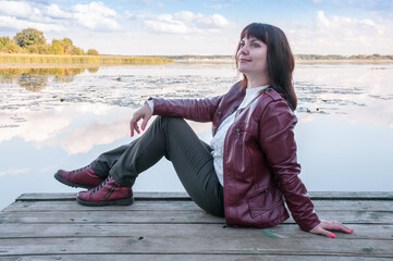 A brunette girl in a white sweater and a red jacket is sitting near the lake in autumn