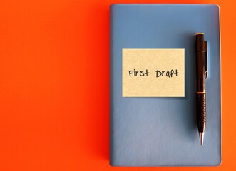 A blue notebook with sicky note written FIRST DRAFT and a black pen isolated on orange background-...