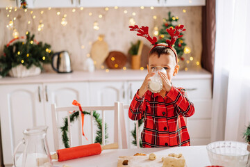 A charming boy in plaid pajamas, drinking milk in a kitchen decorated for Christmas. The concept of comfort and celebration