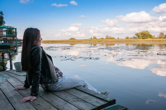 A brunette girl with long hair is sitting on a wooden pier near an autumn lake