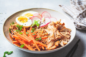 Chicken ramen bowl with carrots, noodles, egg, radish and green onions, gray background.
