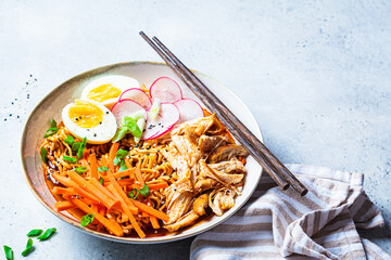 Chicken ramen bowl with carrots, noodles, egg, radish and green onions, gray background.