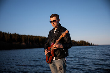 a young guitarist in glasses and a leather jacket plays the electric guitar against the backdrop of nature, a lake and blue sky on the background