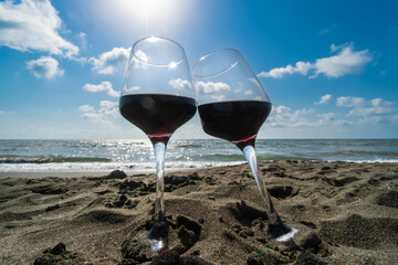 Close-up of two glasses of red wine standing in the sand on the beach against a blue sky with clouds and rays of the sun