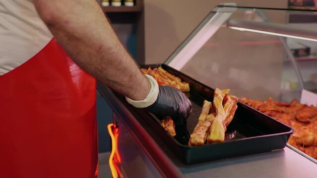 Butchers hands arranging meat on tray for display cabinet at shop. Male placing slices of meat in fulfilling tupperware. Food industry.