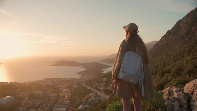 A young woman stands on a viewpoint at sunset and looks down at a coastal town. The tourist destination is Kash, Turkey.