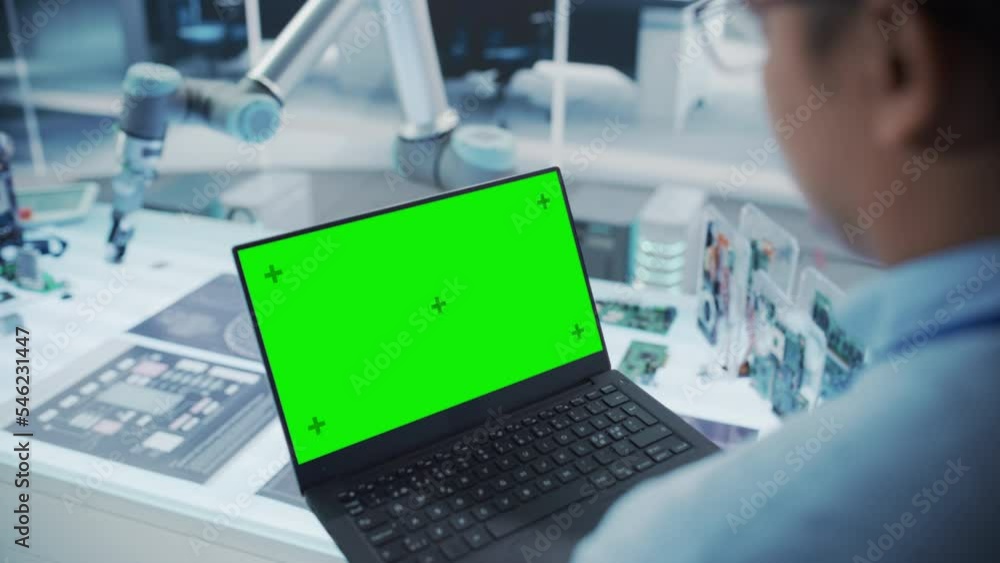 Fototapete Over the Shoulder Shot of a Robotics Engineer Using a Laptop Computer with Green Screen Mock Up Display Screen. Robotic Arm Holding a Microchip. Automation Startup Research and Development Office.
