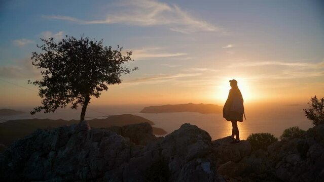 A young woman stands at sunset in the mountains. The silhouette of a girl against the background of the sunset sky and the sea on the top of the mountain. The girl stands on the viewpoint and enjoys t