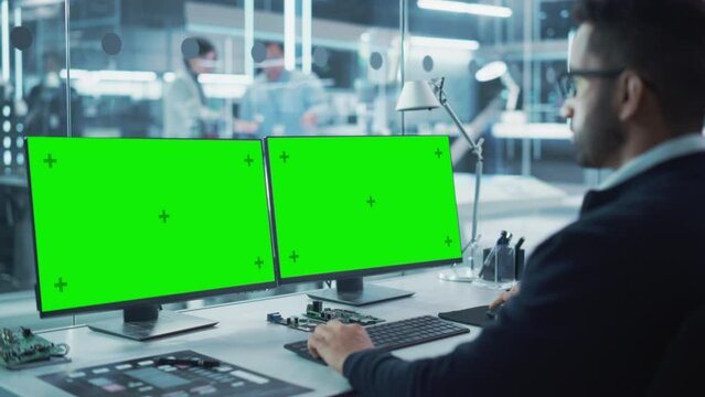 Industrial Engineer Working on a Desktop Computer with Two Green Screen Chromakey Display Screens in a Modern Technological Research and Development Center. Over the Shoulder Shot.