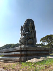Obraz premium A majestic stone sculpture of Lord Ganesha, intricately carved with a large head and crown, holds an axe and modak. Seated against a clear sky and green trees, it reflects rich Hoysala heritage.