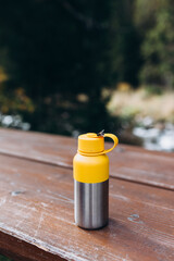 A yellow vacuum theroms stands on a tree desk in the wild. Hiking equipment on footpath in woodland. Travel outdoor concept