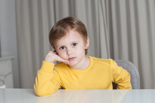 Pensive child boy with brown hair in yellow sweater sitting by the table and looking to the camera on beige curtain background