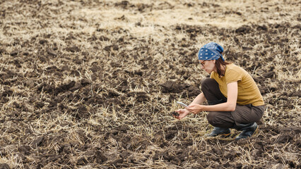 Woman agronomist with a digital tablet sitting on a plowed field with a soil sample in hand,...