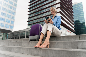 Blond business woman with travel suitcase typing on cellphone sitting outside office building ready...