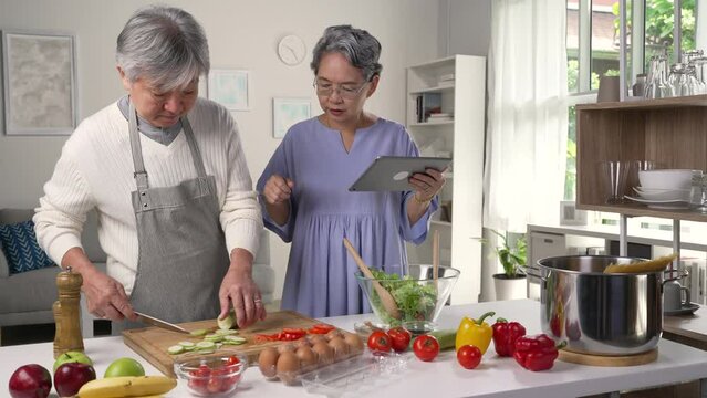 Happy Asian senior couple is using a digital tablet, talking and smiling while cooking together in kitchen.